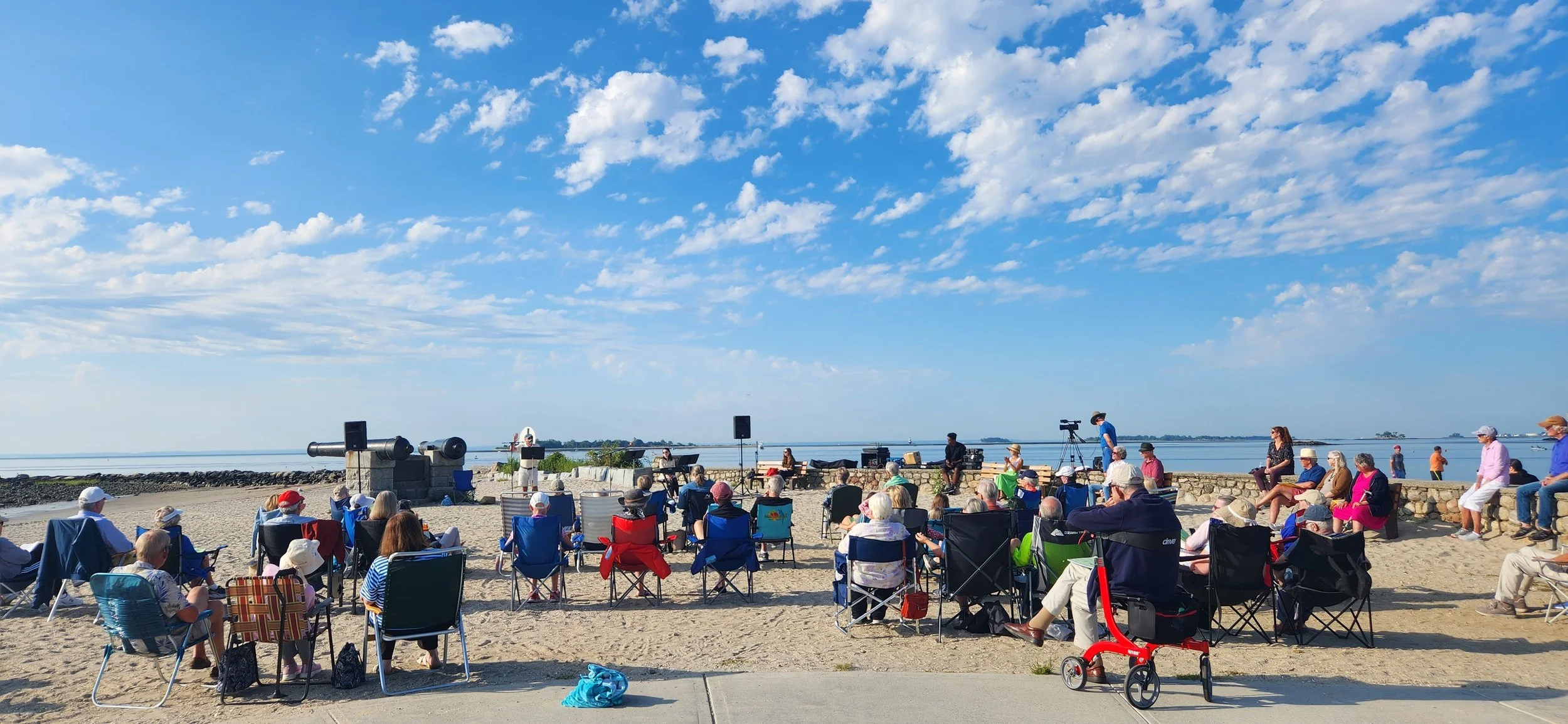 A gathering of many people in beach chairs on the sand facing the ocean while a pastor preaches