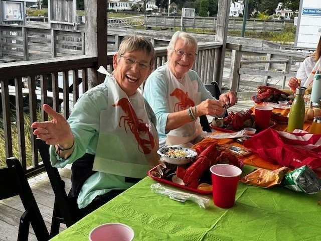 Two women sitting at a table outside, enjoying a meal with lobster and sides, smiling and waving at the camera.