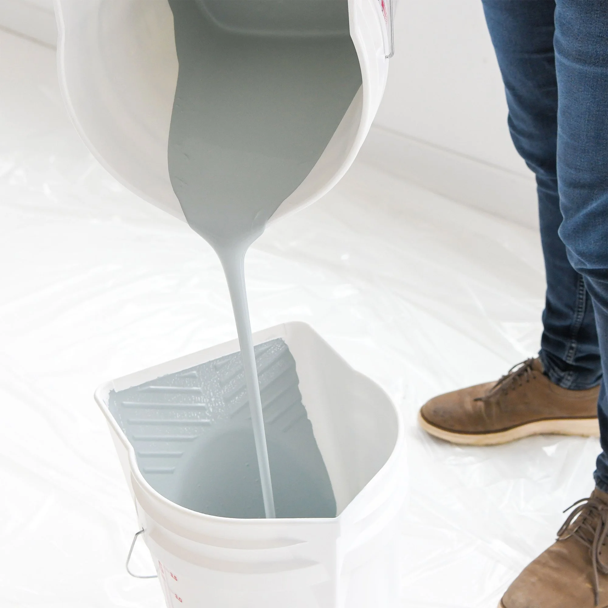 Pouring gray paint from a bucket into a paint tray, with a person in jeans and brown boots standing nearby.