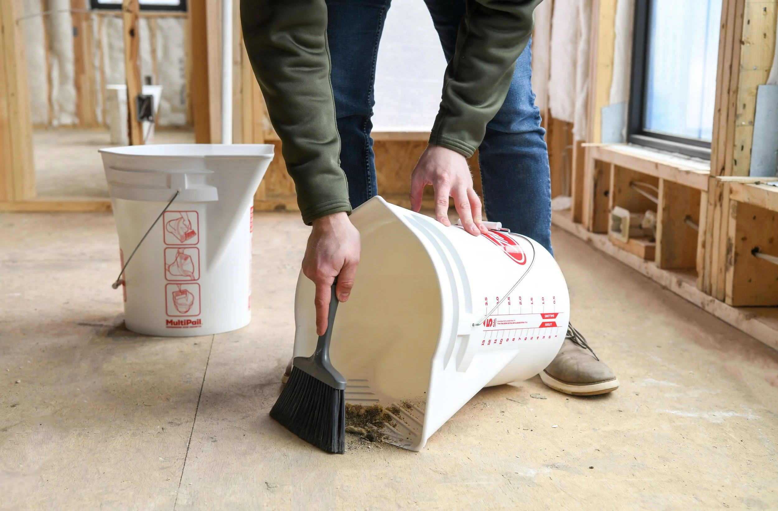 Person sweeping up dirt in an unfinished room with exposed wooden framing.