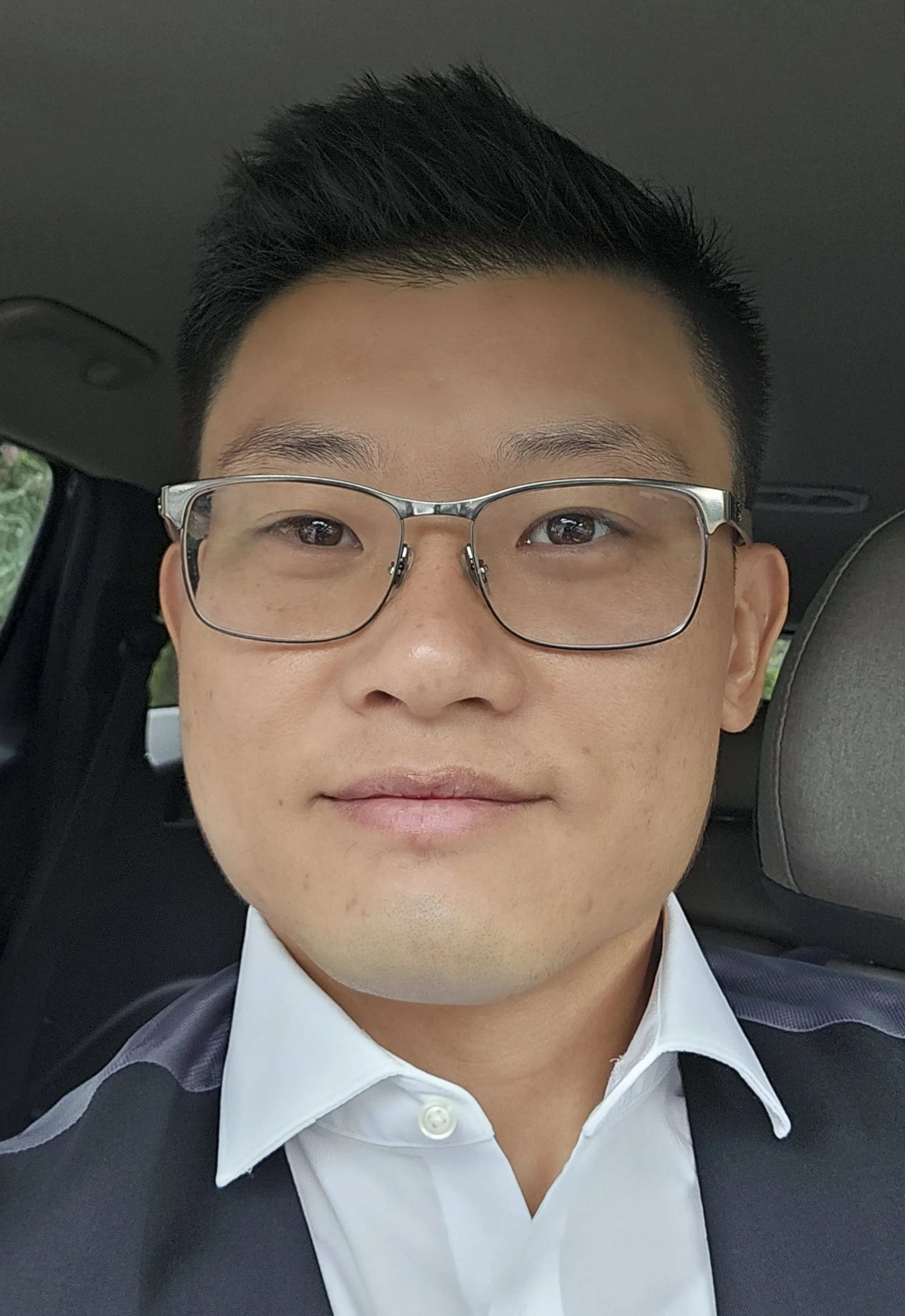 Close-up of a young man with short black hair and glasses, smiling inside a car, wearing a white shirt and dark jacket.