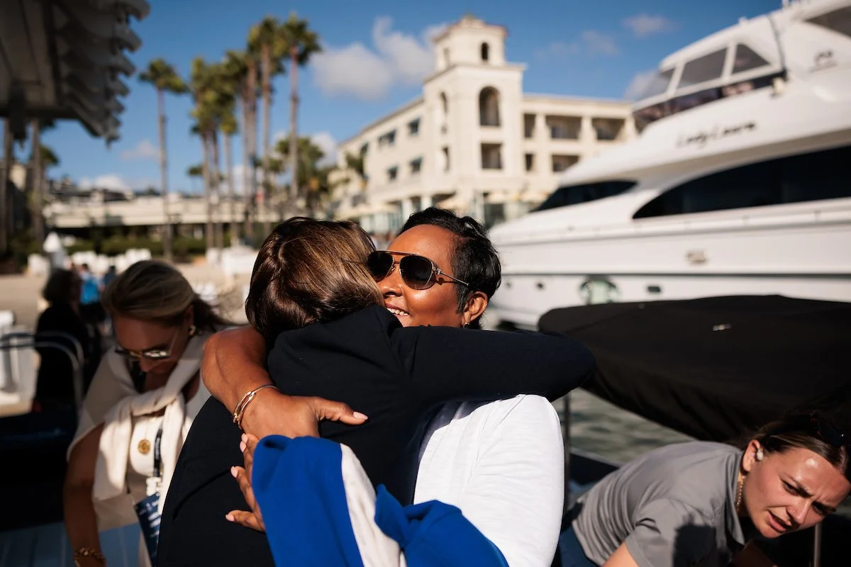 Two women hugging with smiles, one wearing sunglasses, in front of a yacht docked at a marina, with a building and palm trees in the background.