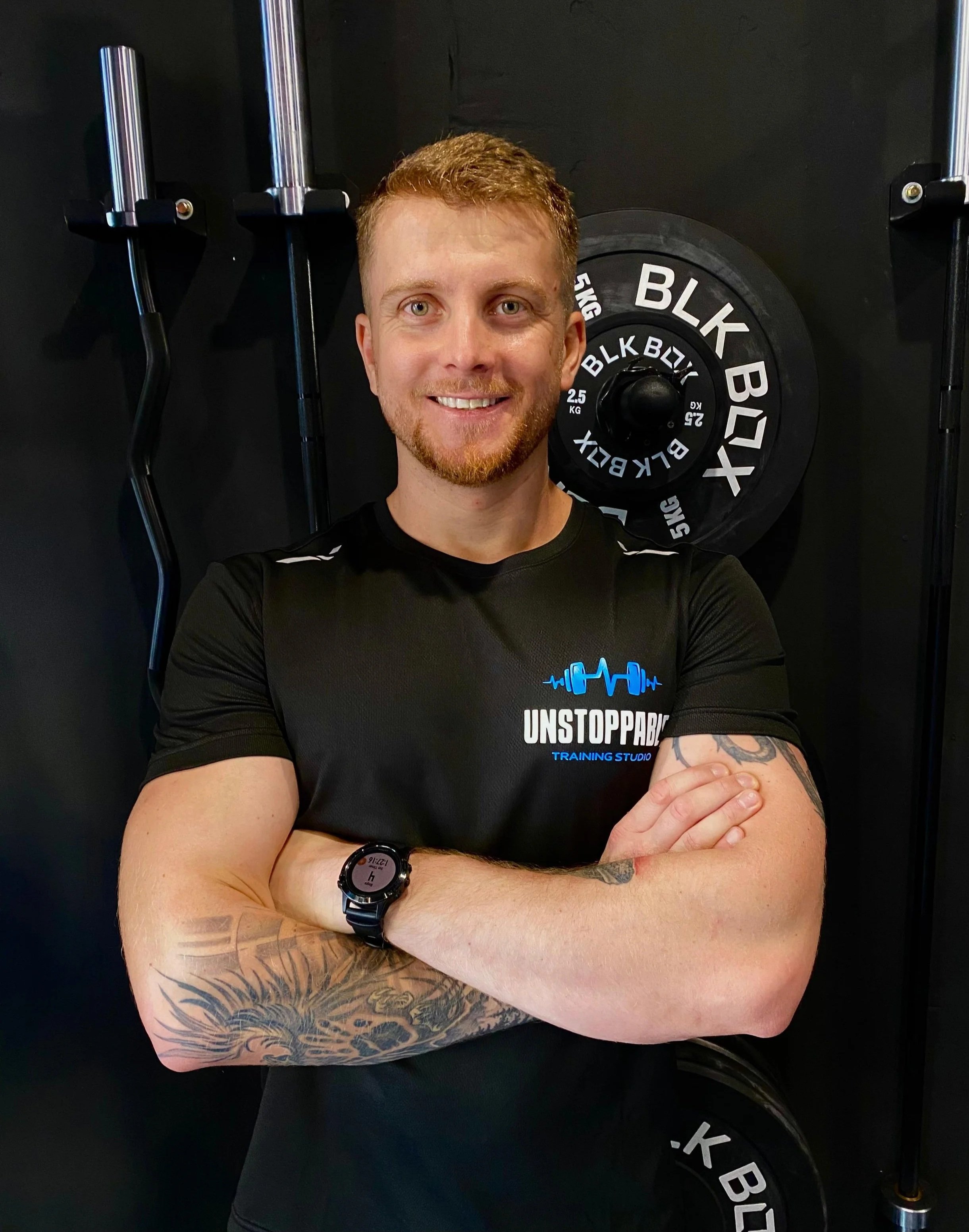 Smiling man with tattoos on his arms standing with arms crossed in front of weights at a gym, wearing a black T-shirt with "UNSTOPPABLE TRAINING STUDIO" logo.