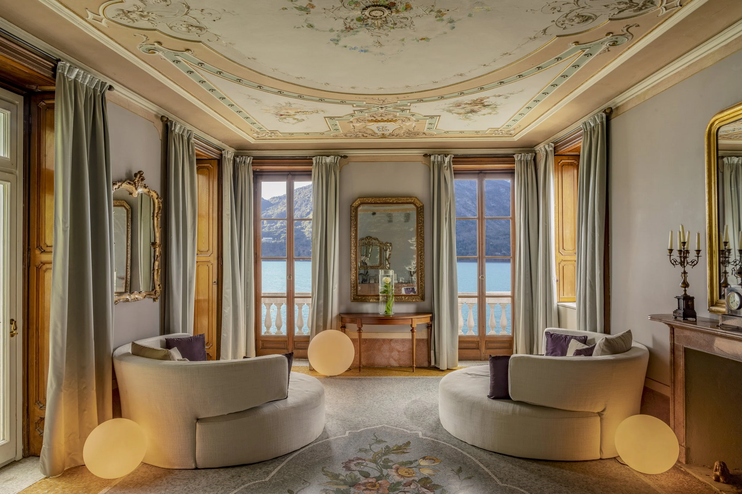 Elegant living room with large windows overlooking a lake and mountains, featuring cream-colored curved sofas, gold-framed mirrors, and soft lighting.