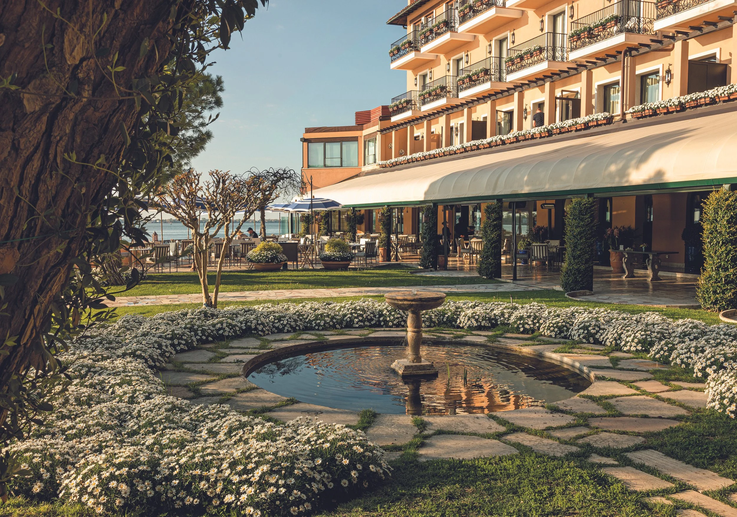 Outdoor view of a hotel or resort with a garden, fountain, patio seating, and a multi-story building in the background.