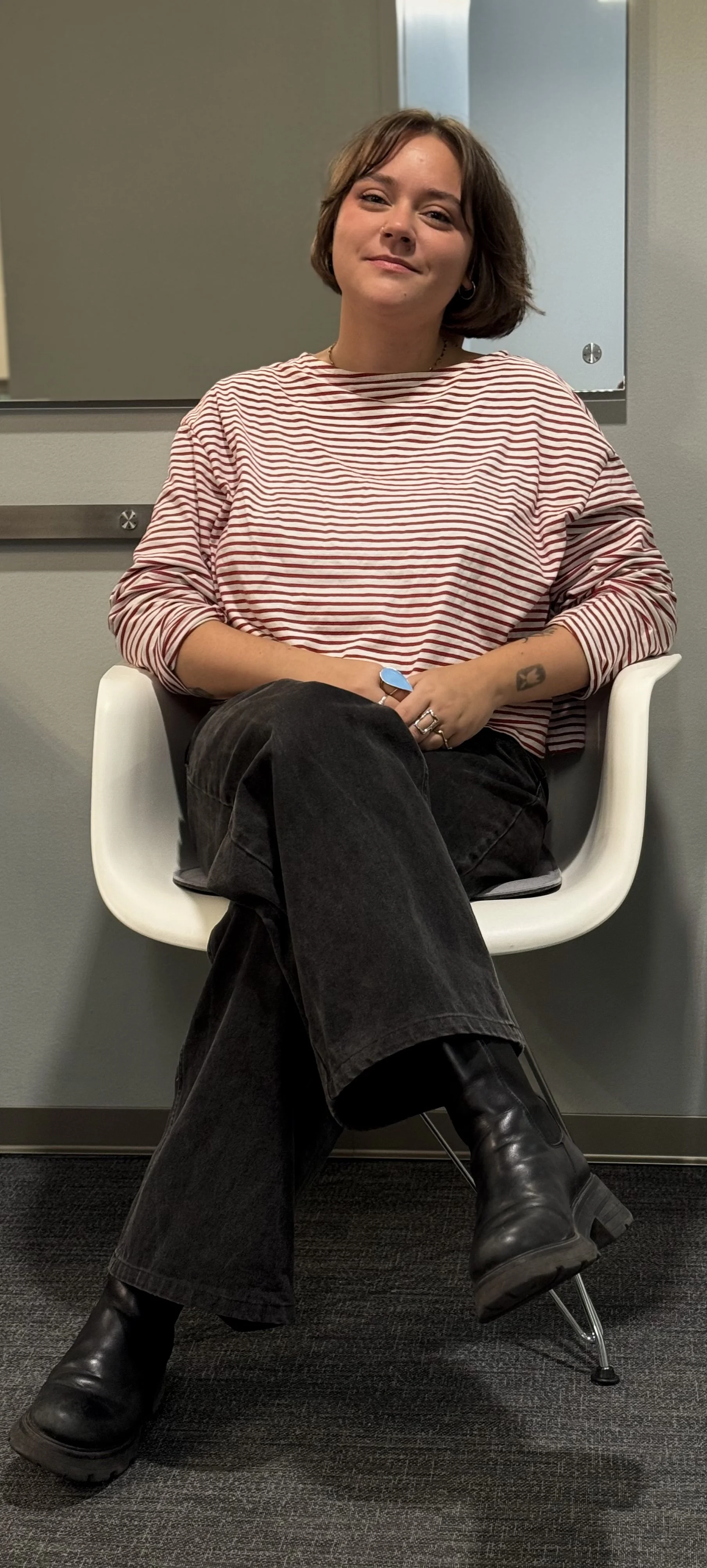 Eine junge Frau sitzt in einem weißen Stuhl vor einem grauen Hintergrund, trägt eine rot-weiße gestreifte Bluse, schwarze Hosen und schwarze Stiefel. Sie schaut in die Kamera mit einem leichten Lächeln.
