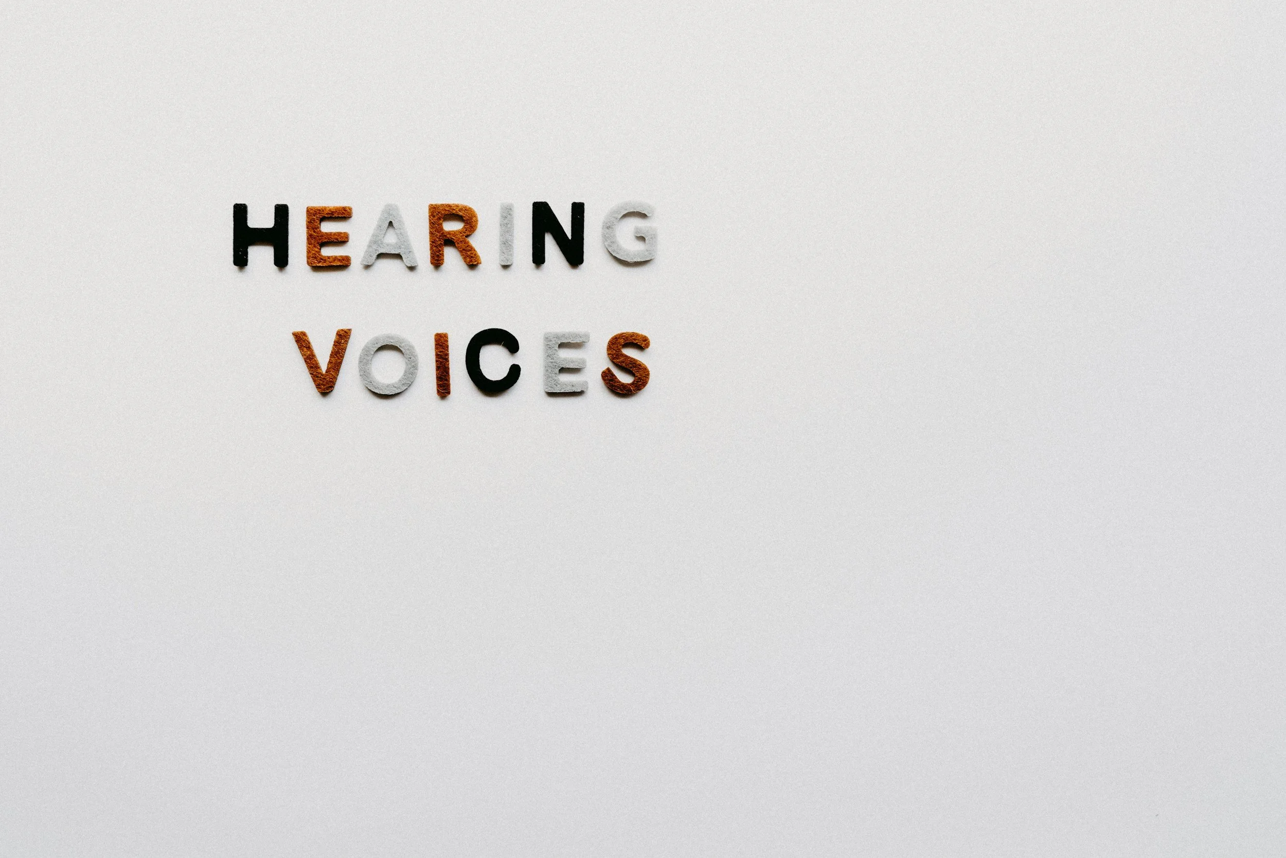 Buchstaben auf weißem Hintergrund, die das Wort "HEARING VOICES" bilden, mit verschiedenen Farben und Texturen.