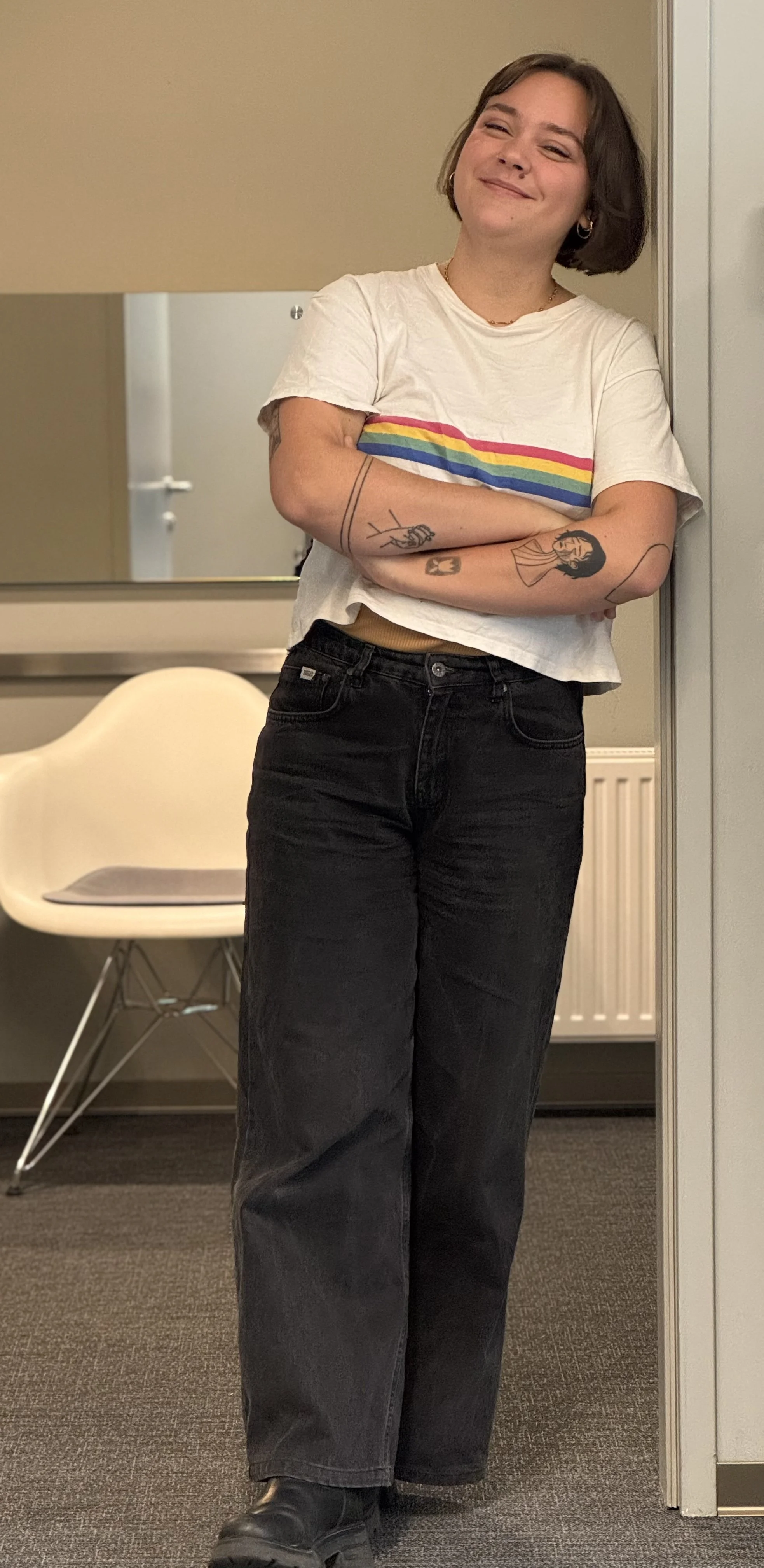 Junge Frau mit kurzen braunen Haaren, Tatoos auf den Armen, trägt ein weißes T-Shirt mit Regenbogen-Streifen, schwarze Jeans und schwarze Schuhe, steht lächelnd in einem Raum, lehnt an der Wand.