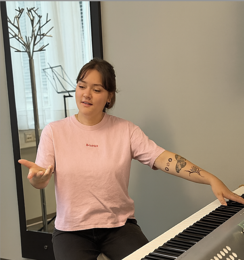 Eine junge Frau spielt Klavier und wirkt dabei nachdenklich, trägt ein rosa T-Shirt mit der Aufschrift 'Bisous' und hat Tattoos am linken Arm.