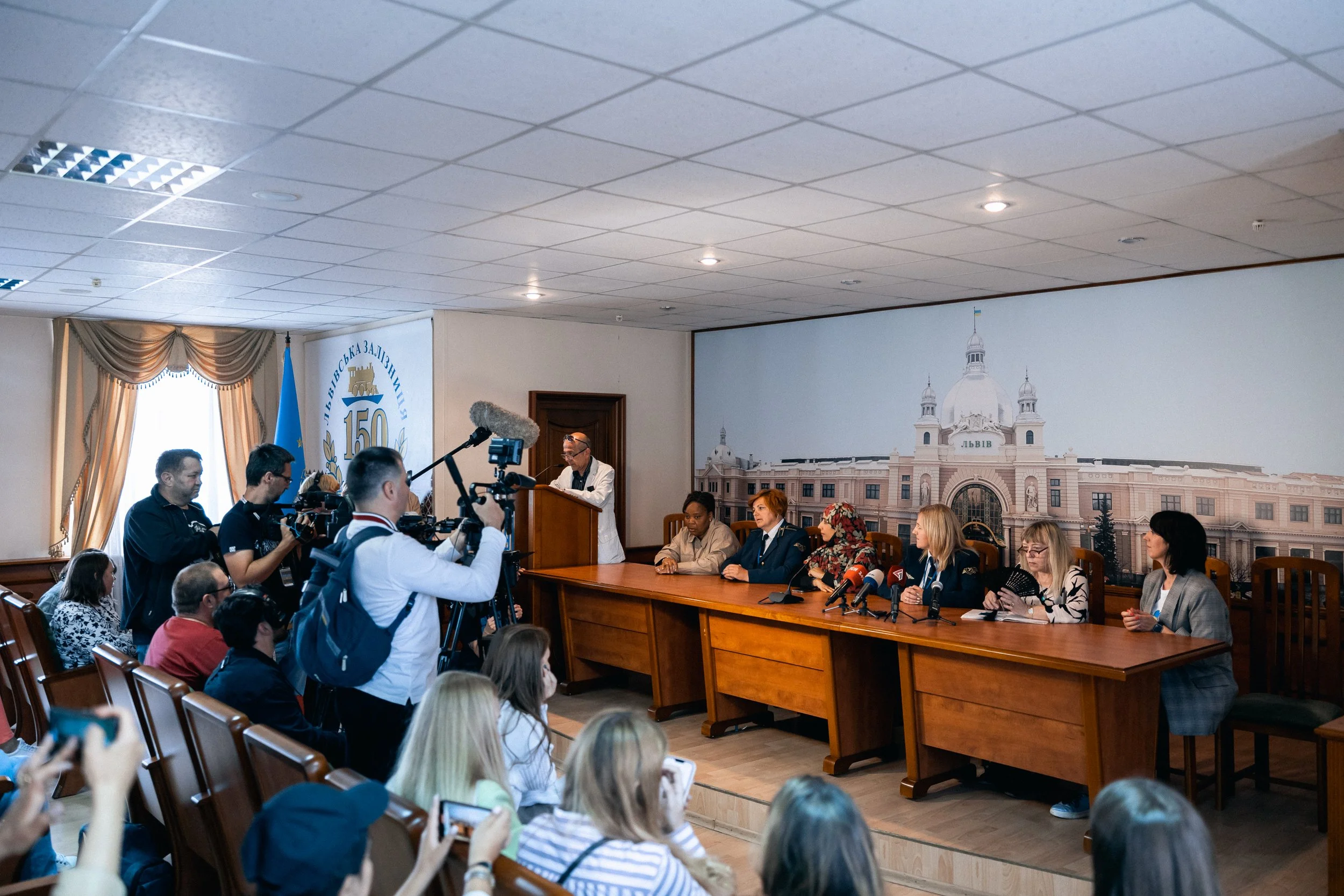 Press conference with six women seated at a long table and several journalists and photographers capturing the event, behind them a large wall mural of a historical building with domes.