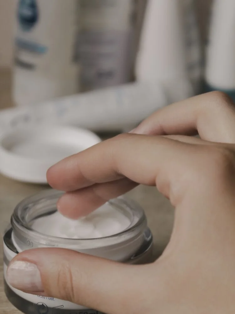 Hand dipping finger into jar of white cream, with skincare or cosmetic bottles in the background.