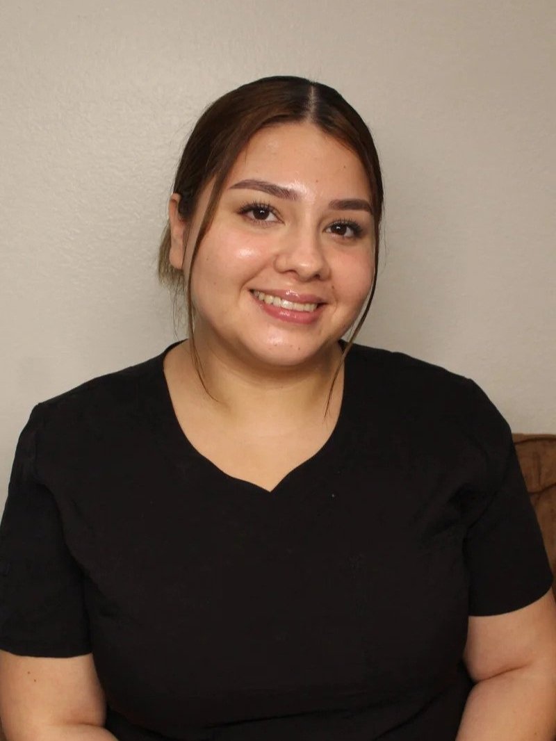 A woman with light brown hair in a black shirt smiling at the camera against a plain wall.