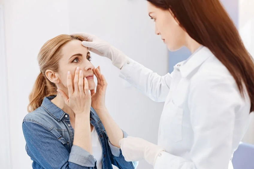 A woman in a denim shirt has her face examined by a female doctor in a white coat in a clinical setting.