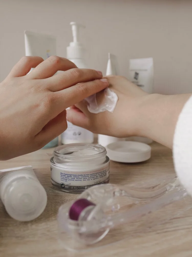 A person applying a white cream or moisturizer to their hand using a cotton pad, with various skincare products like jars and tubes on a wooden surface, in a bathroom or skincare setting.