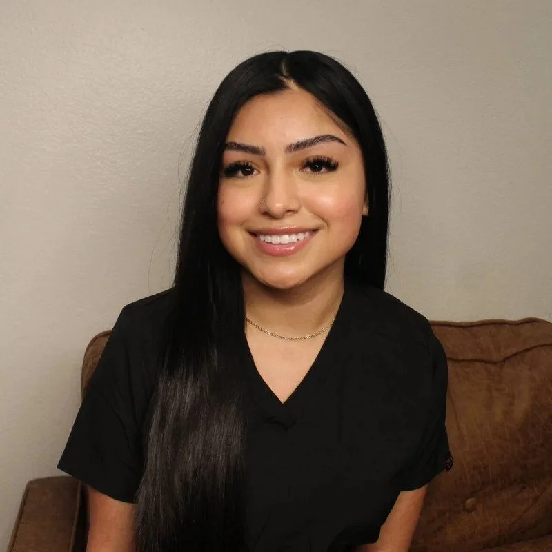 A young woman with long black hair, smiling, sitting on a brown couch against a plain beige wall.