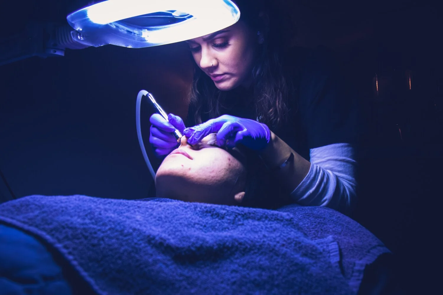 A woman performing a cosmetic tattoo procedure on a person's eyebrow using a tattoo machine, under a bright light, with both wearing gloves.