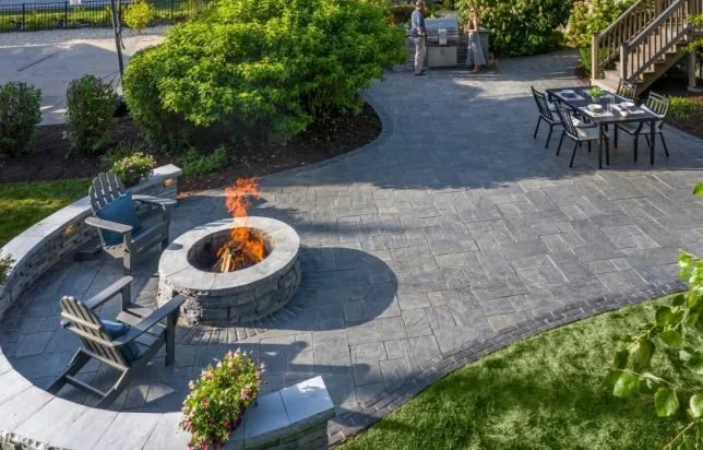 An outdoor patio with seating, a fire pit, greenery, and a person grilling in the background.