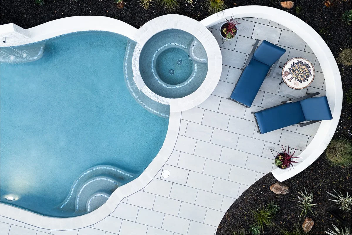 A swimming pool with a circular hot tub attached, surrounded by a white patio with two blue lounge chairs, a small round table, and potted plants.