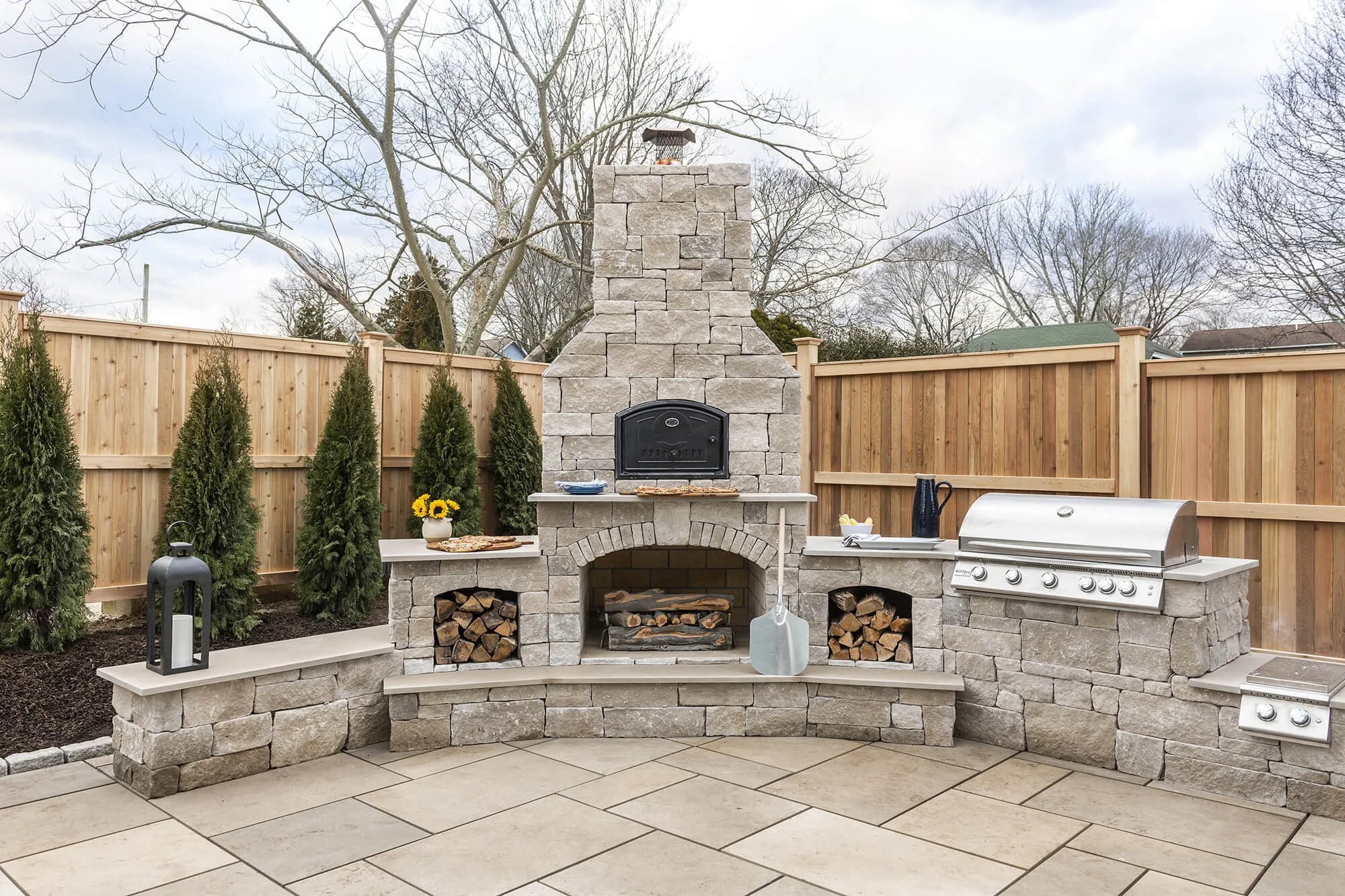 Outdoor patio with a stone fireplace, wood storage, and a stainless steel grill, enclosed by a wooden fence with trees in the background.