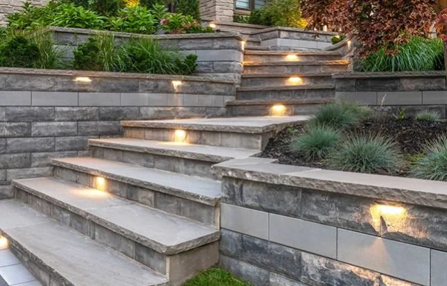 Outdoor stone staircase with built-in lighting, leading up through landscaped garden beds with green plants and bushes.