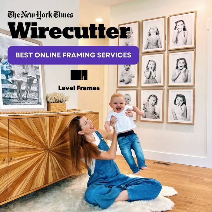 Mother playing with her baby in a bright modern room featuring a gallery wall of framed black-and-white portraits, with The New York Times Wirecutter badge naming Level Frames as Best Online Framing Services