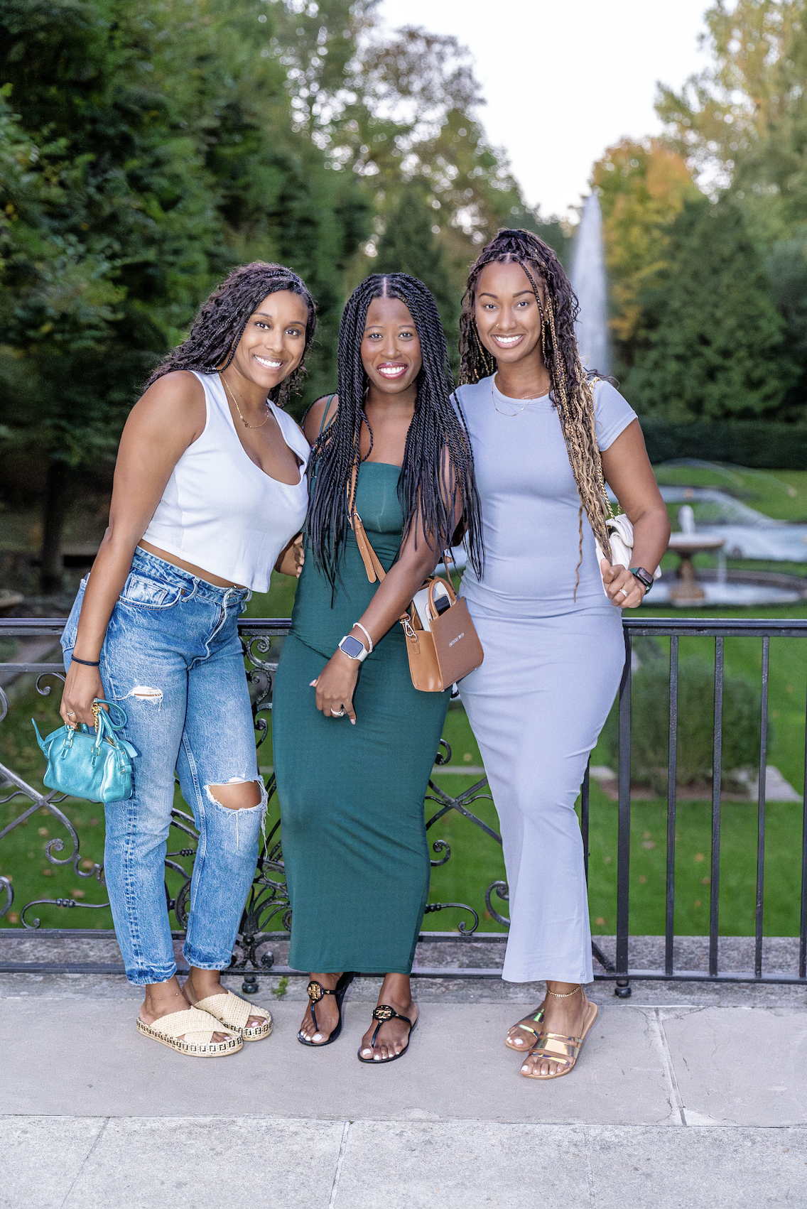 Three women standing outdoors on a balcony with green trees and a fountain in the background, smiling at the camera.