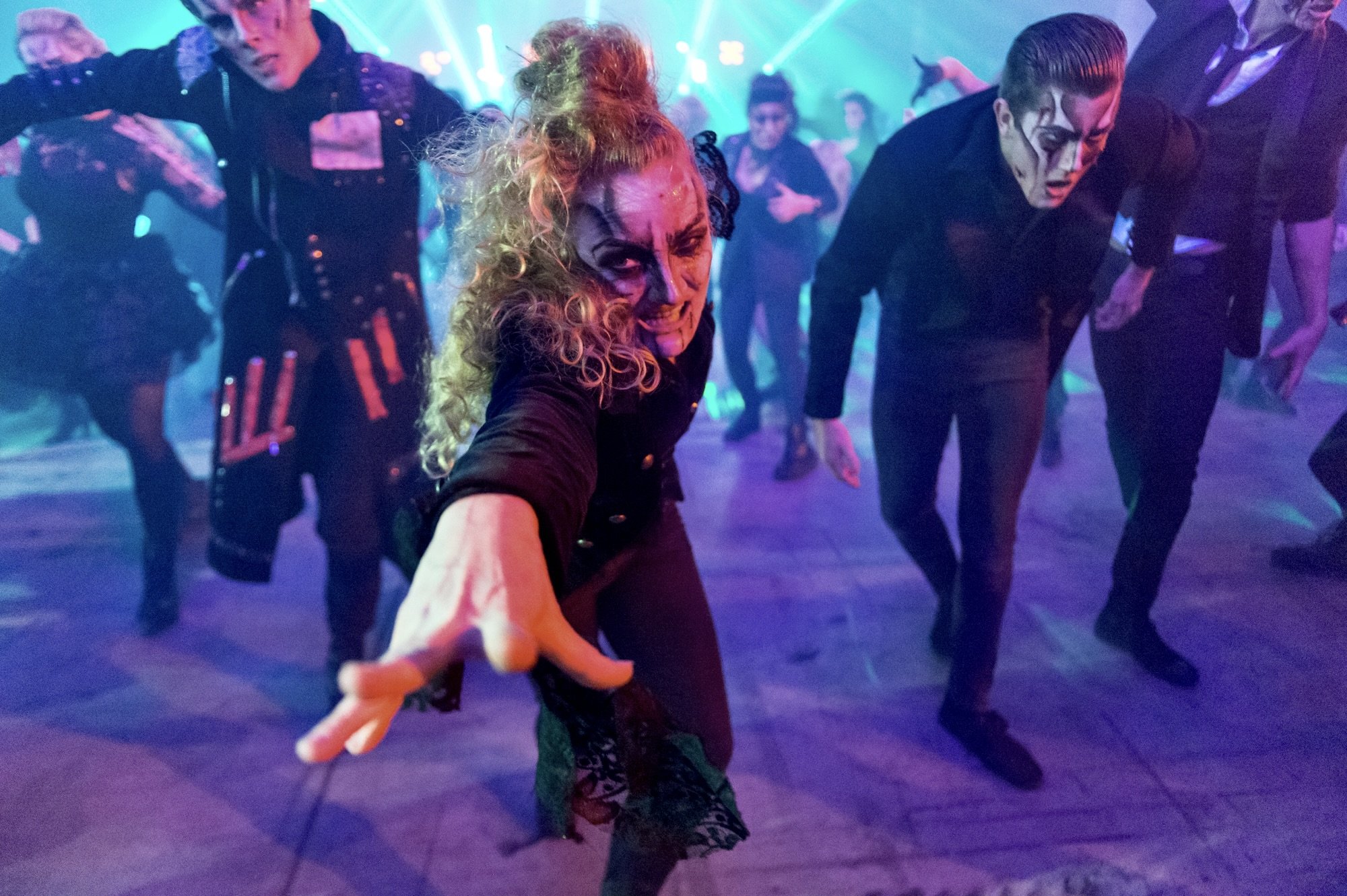 People dancing at a party with colorful lighting, with a woman with curly blonde hair and face makeup reaching towards the camera.