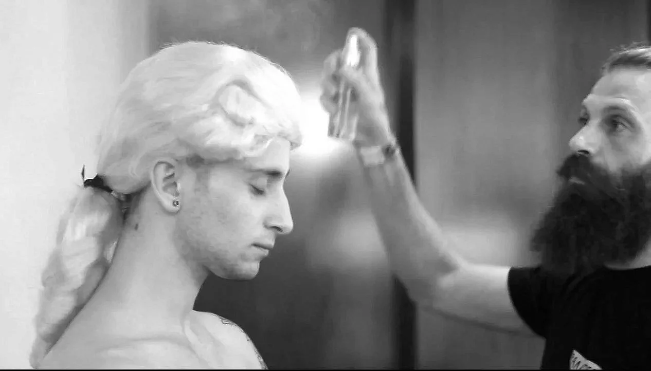 A person with a large beard and short hair is painting a woman's face with a paintbrush, in black and white.