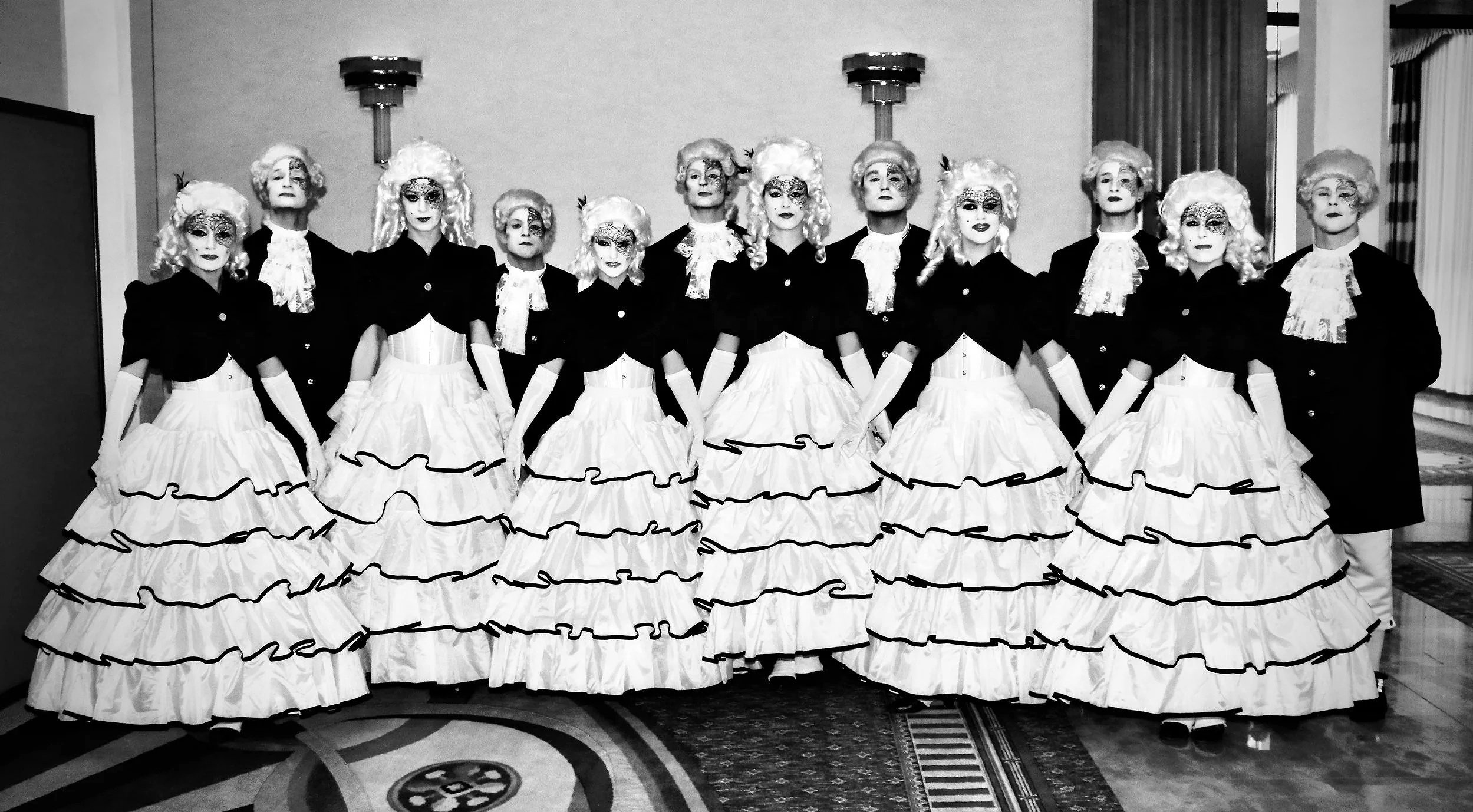 A group of eleven women dressed in historical costumes, with long, ruffled white skirts, dark jackets, and lace-trimmed blouses, standing in a line indoors, wearing wigs and makeup, likely for a performance or costume event.