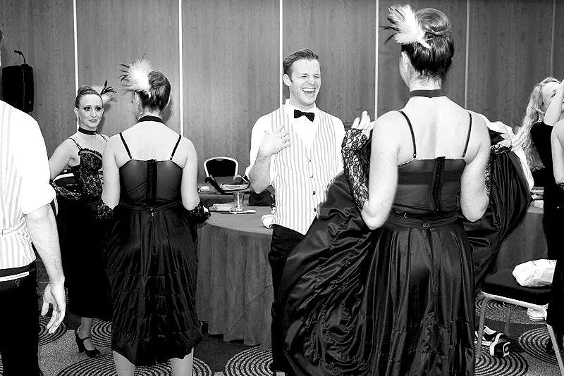People laughing and talking at a social event or party, with some women in elaborate dresses and one man in a tuxedo with a bowtie.