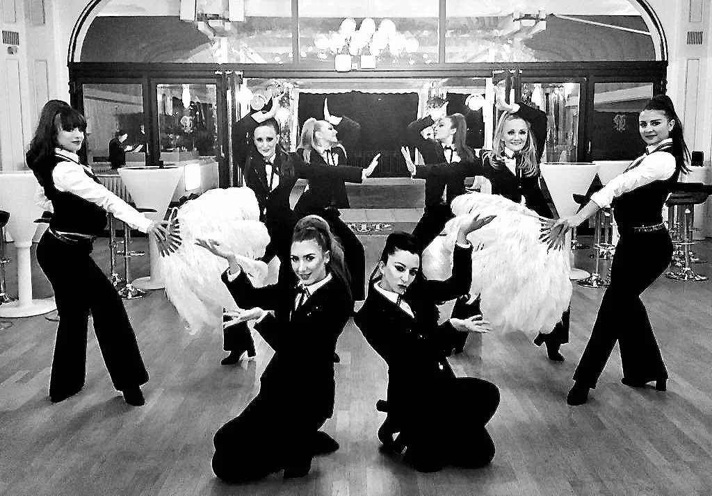 Group of seven women in formal attire, some holding large feathered fans, posing in a dance formation indoors with a bar in the background.