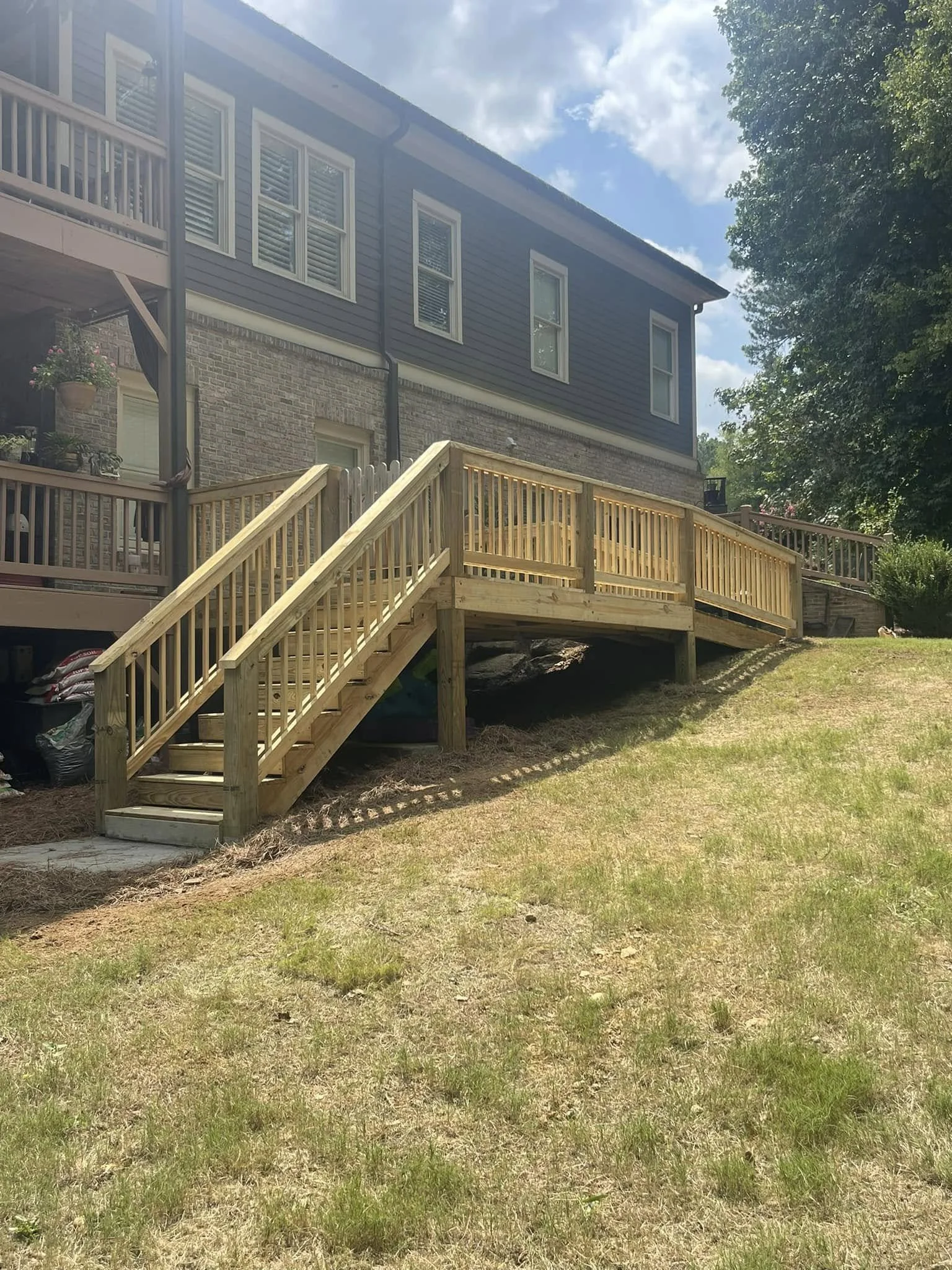 Wooden stairway and ramp leading to the backyard of a two-story house with gray siding and brick exterior. The house has multiple windows, a porch, and hanging flower baskets. The yard has grass and some trees.