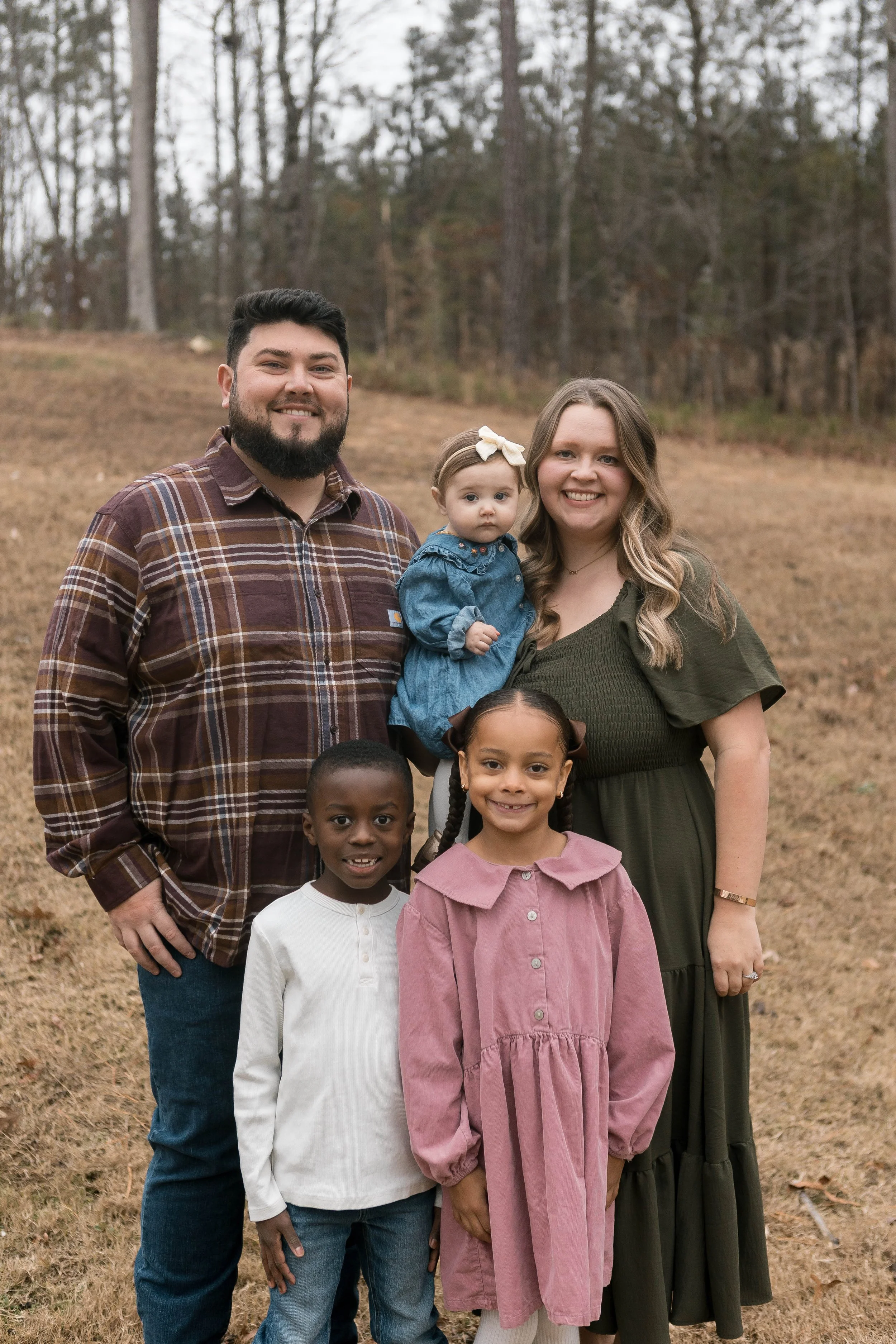 Family photo outdoors with two adults and three children, standing on a grassy field with trees in the background.