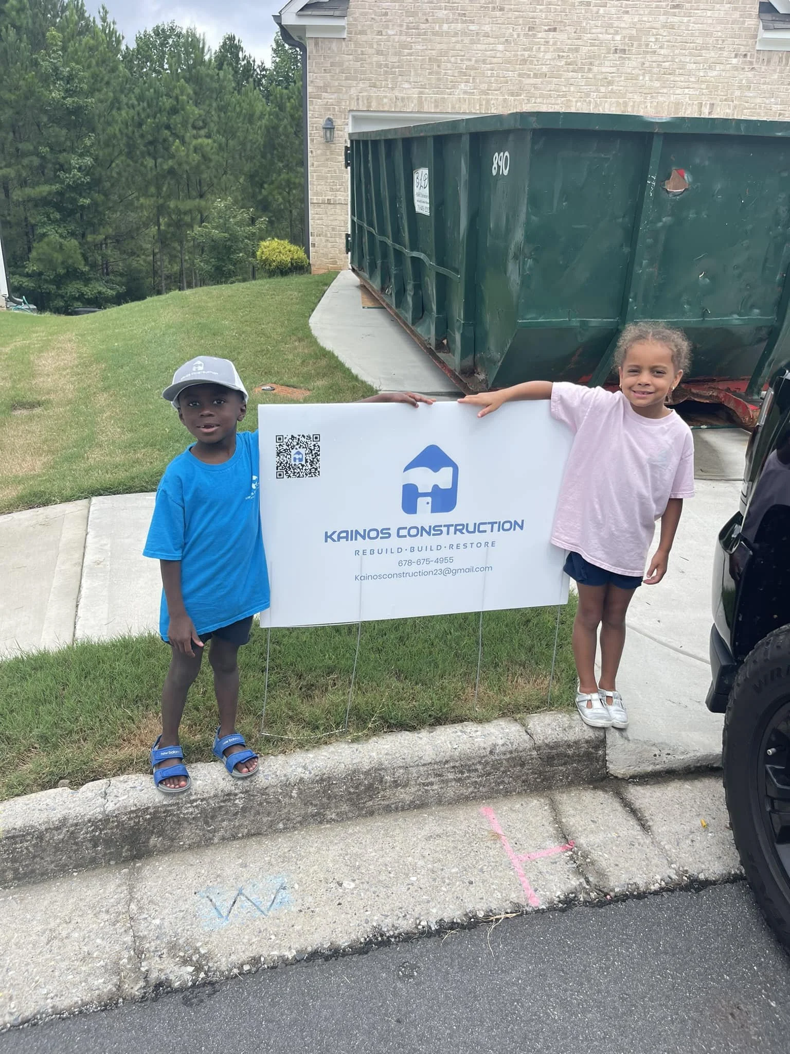 Two children, a boy and a girl, standing next to a sign for Kainos Construction in front of a house with a large green dumpster in the driveway. The boy is wearing a blue shirt, gray shorts, a white cap, and sandals. The girl is wearing a pink shirt, dark shorts, and white shoes. They are outdoors on a concrete sidewalk and grass, with trees and a house in the background.