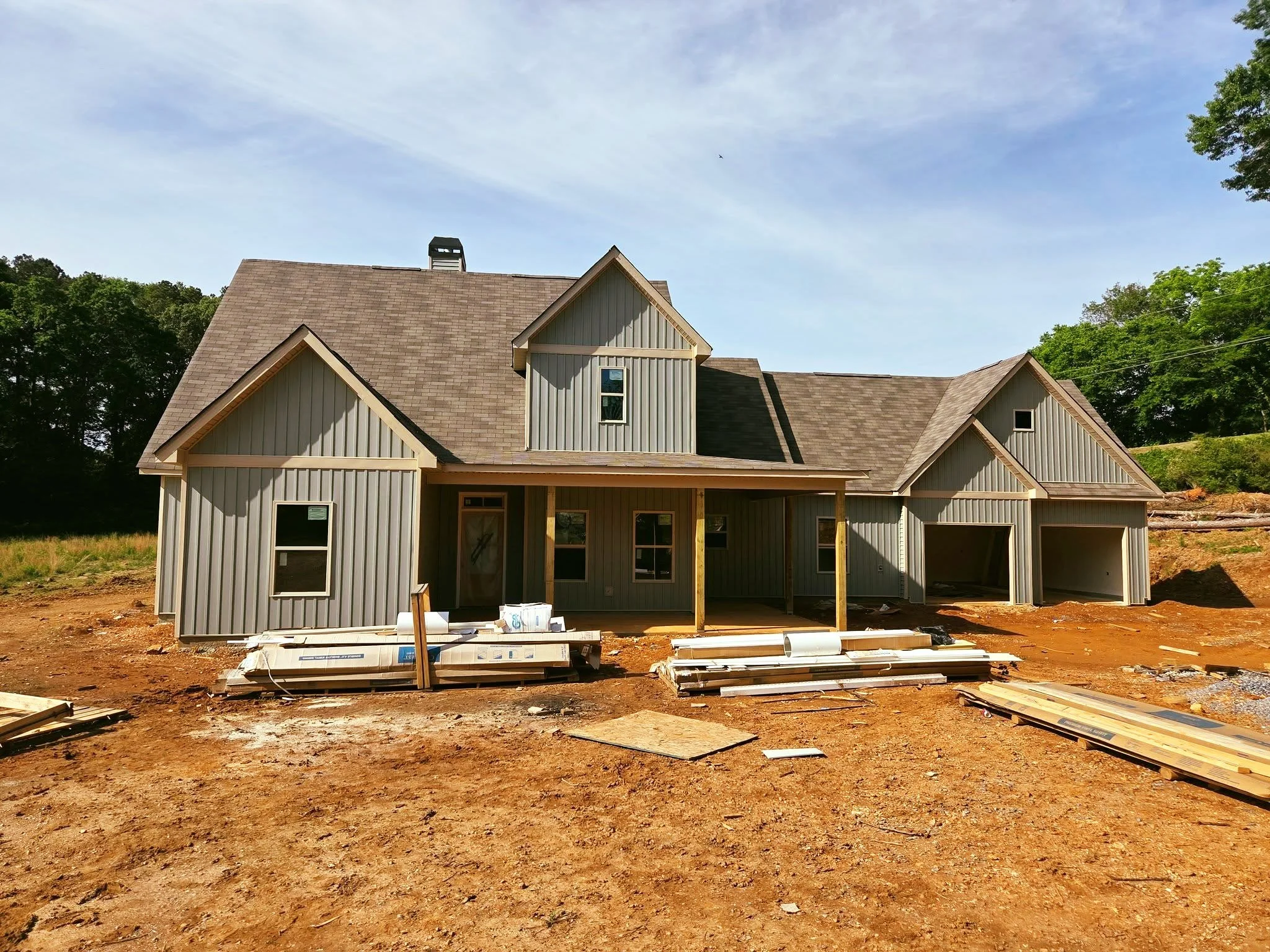 Under construction house with gray siding, multiple gables, and a covered porch, surrounded by dirt and construction materials, with trees and a blue sky in the background.