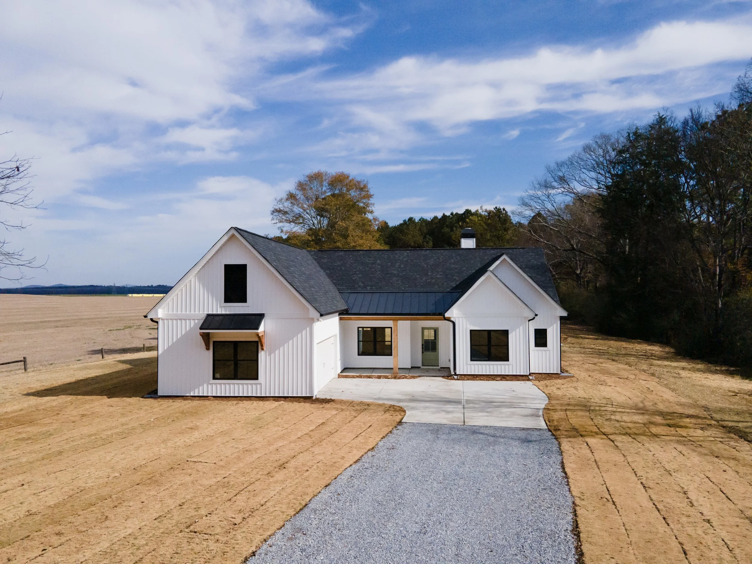 A new white house with black roof shingles is situated on a large plot of land with bare patches of grass and dirt, and a gravel driveway leading to the front door, surrounded by trees under a partly cloudy sky.