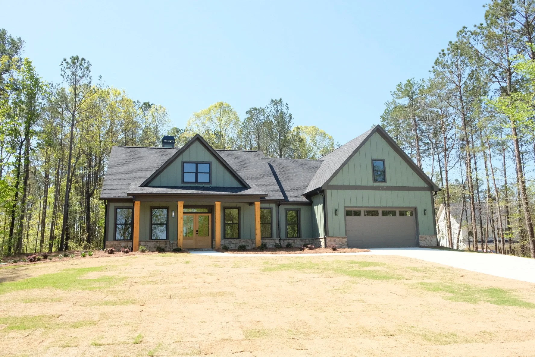 Newly built house with green exterior, black roof, front porch with wooden columns, and attached garage, surrounded by trees under a clear blue sky.