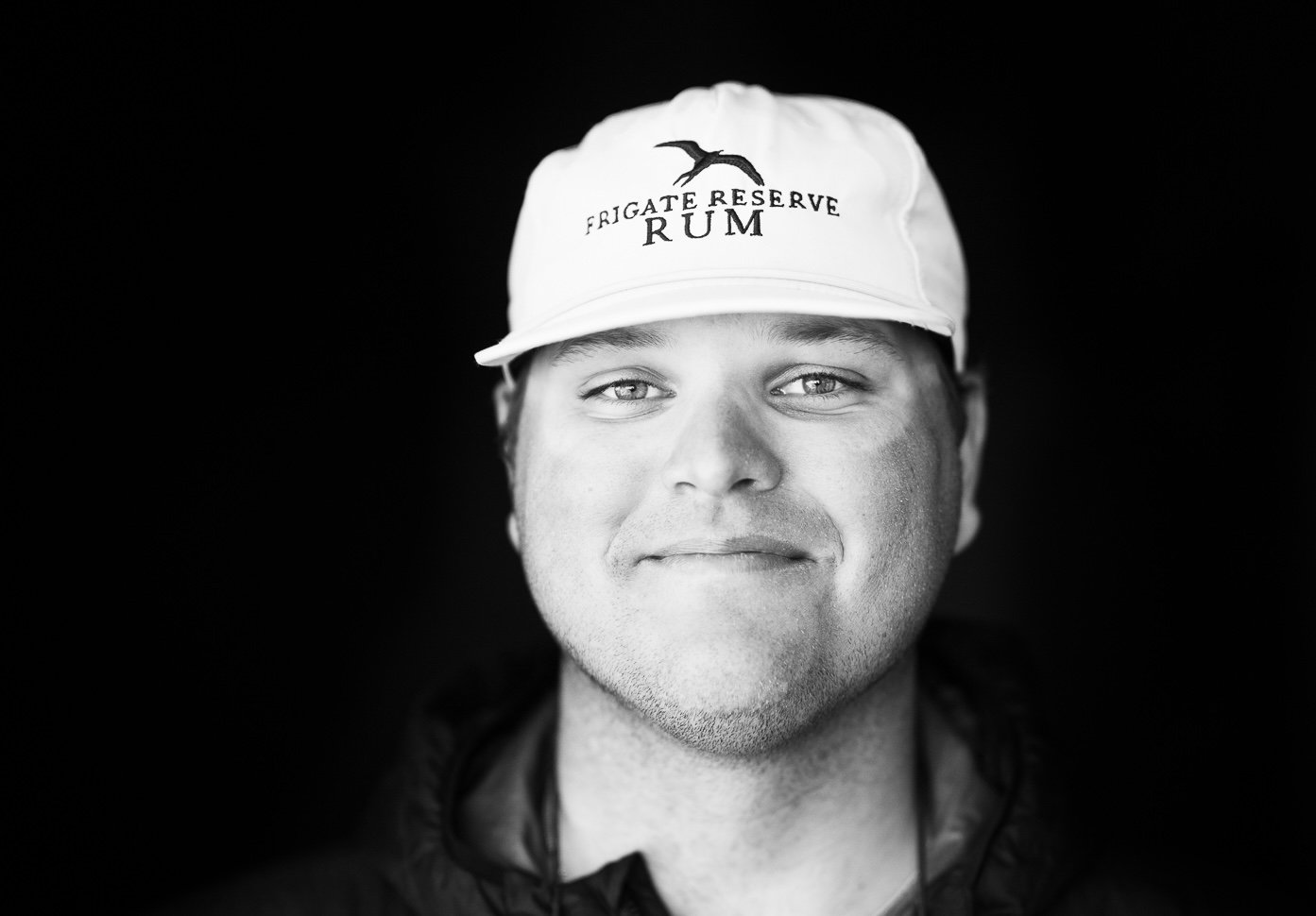 Black and white portrait of a smiling man wearing a white cap with a bird logo and the words "Fragate Reserve Rum".
