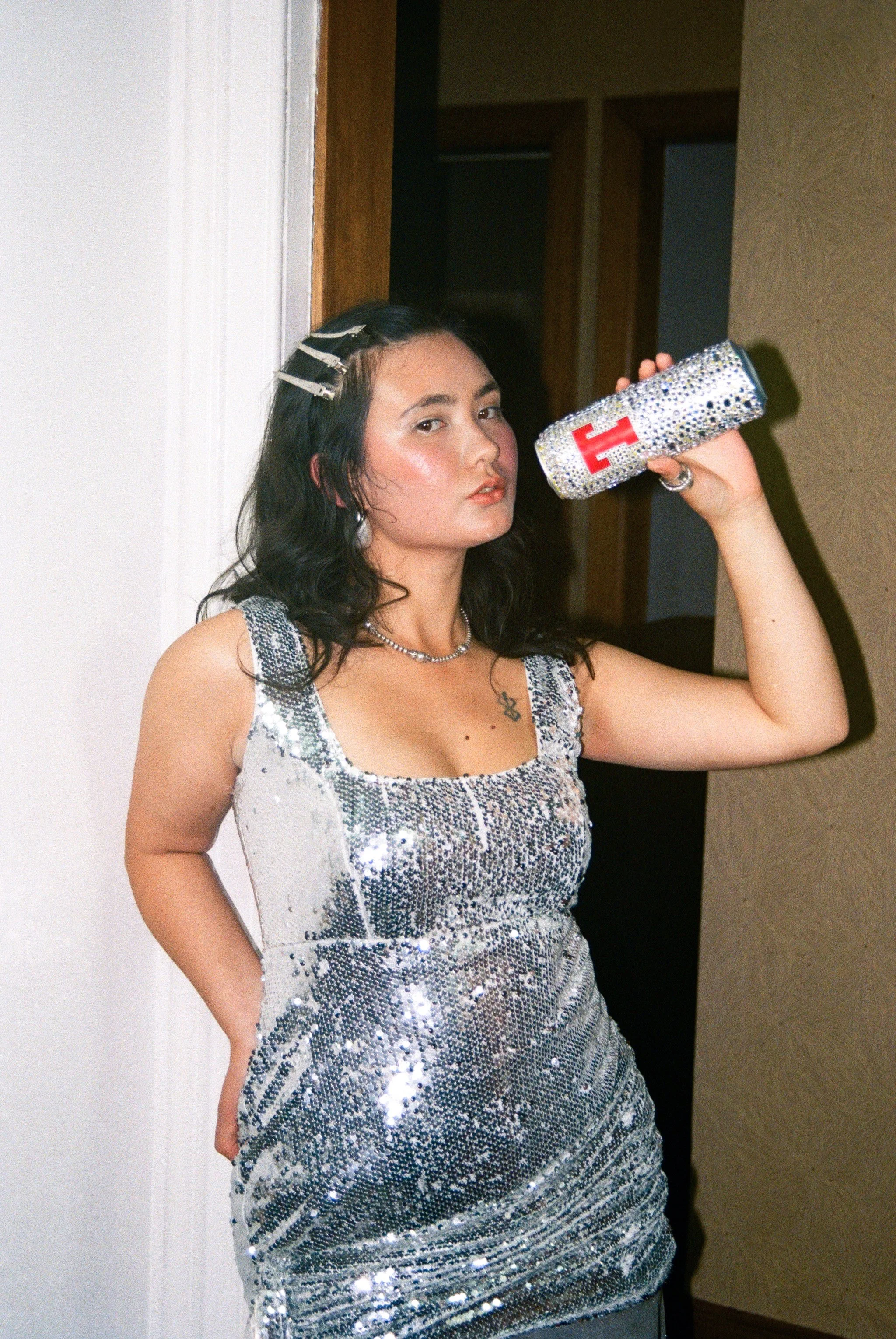 A woman with dark hair styled with hair clips, wearing a silver, sequined dress, holding a matching sparkly clutch, standing indoors next to a doorframe.