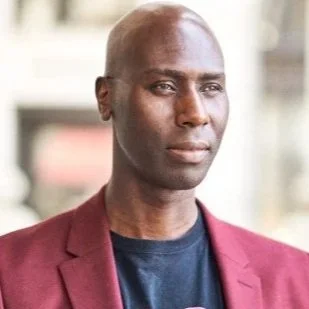 A man with a bald head wearing a maroon blazer and dark shirt, standing outdoors.