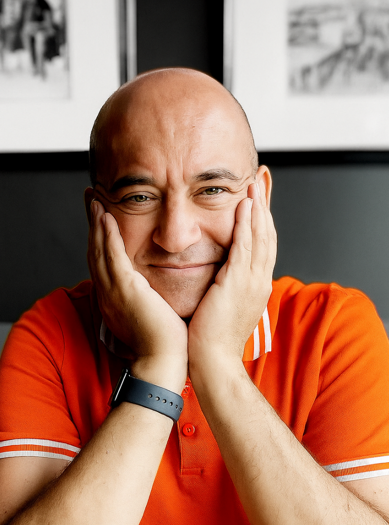 Smiling man with bald head, wearing an orange polo shirt and a black wristband, resting his face in his hands.