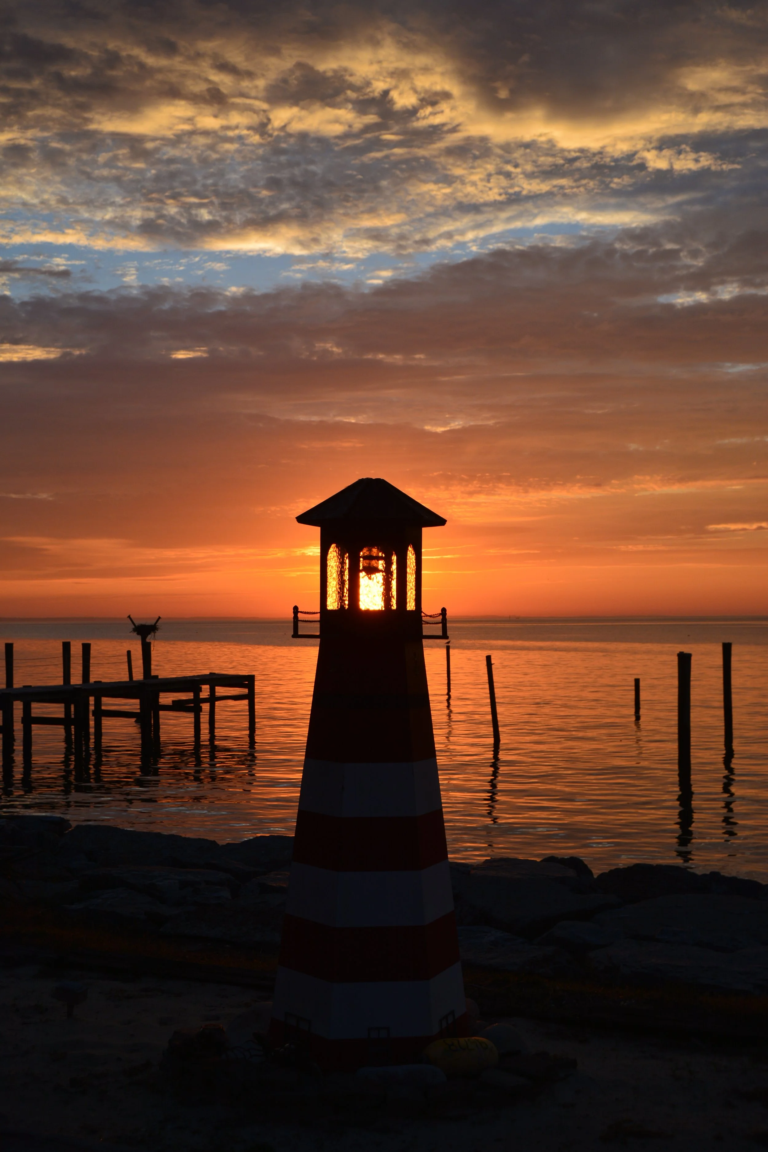 Silhouette of a lighthouse on a beach during sunset, with a wooden pier extending into the water and clouds in the sky.