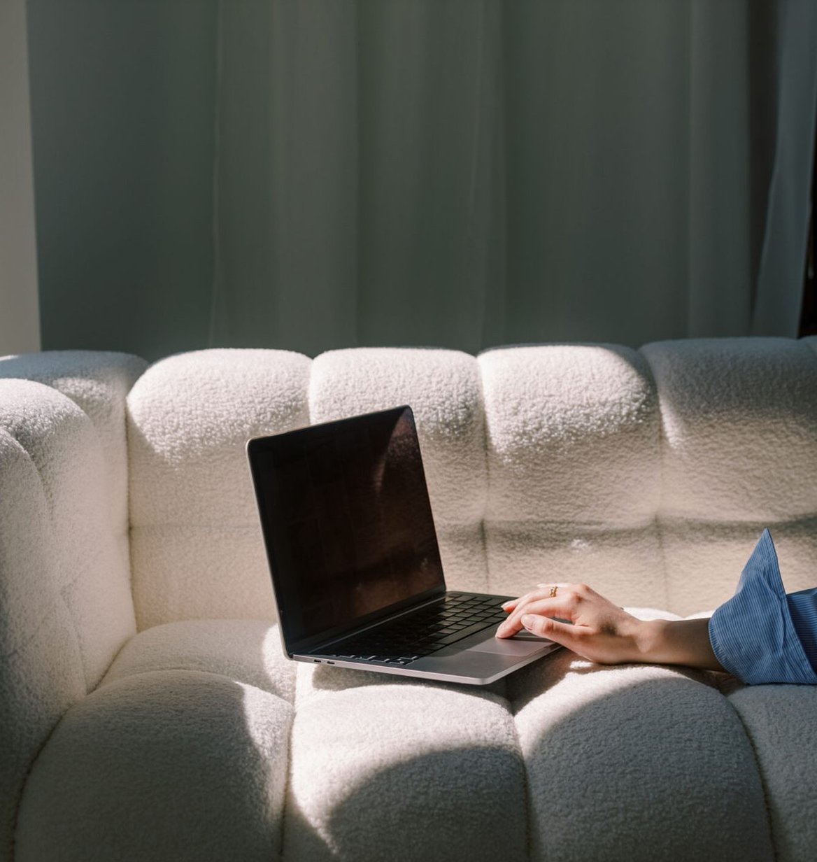 A person resting their hand on a laptop on a white upholstered sofa, with green curtains in the background.