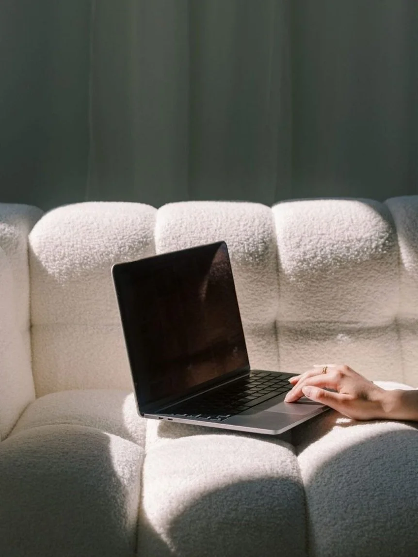 A person using a laptop on a textured cream-colored sofa with a dark curtain in the background.