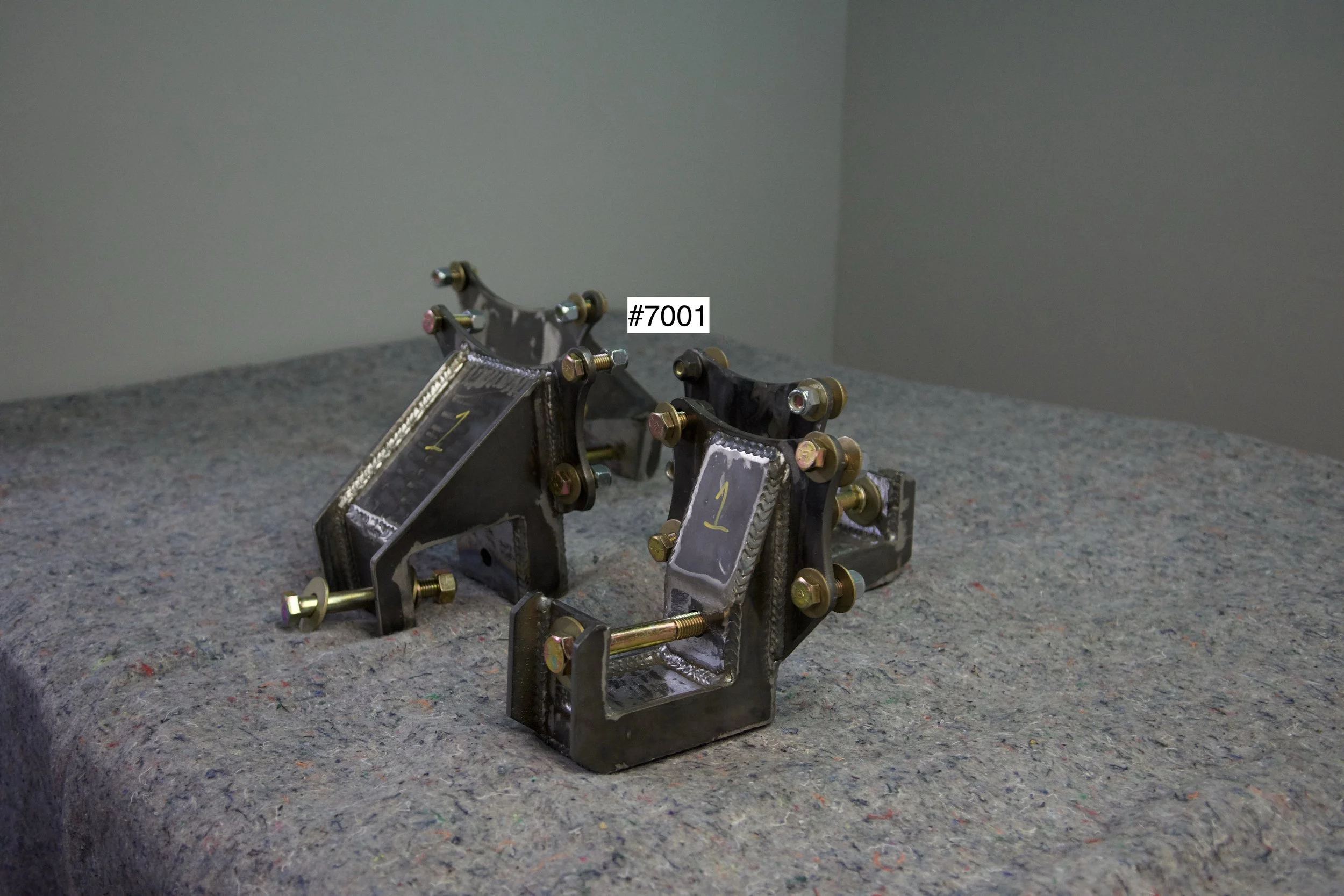 SuperBronco Rear Spring Mounts Rev. 1 – raw bare steel brackets shown from front and side angles, part numbers #7001 and #7002. Fabricated mounts for Bronco suspension builds.
