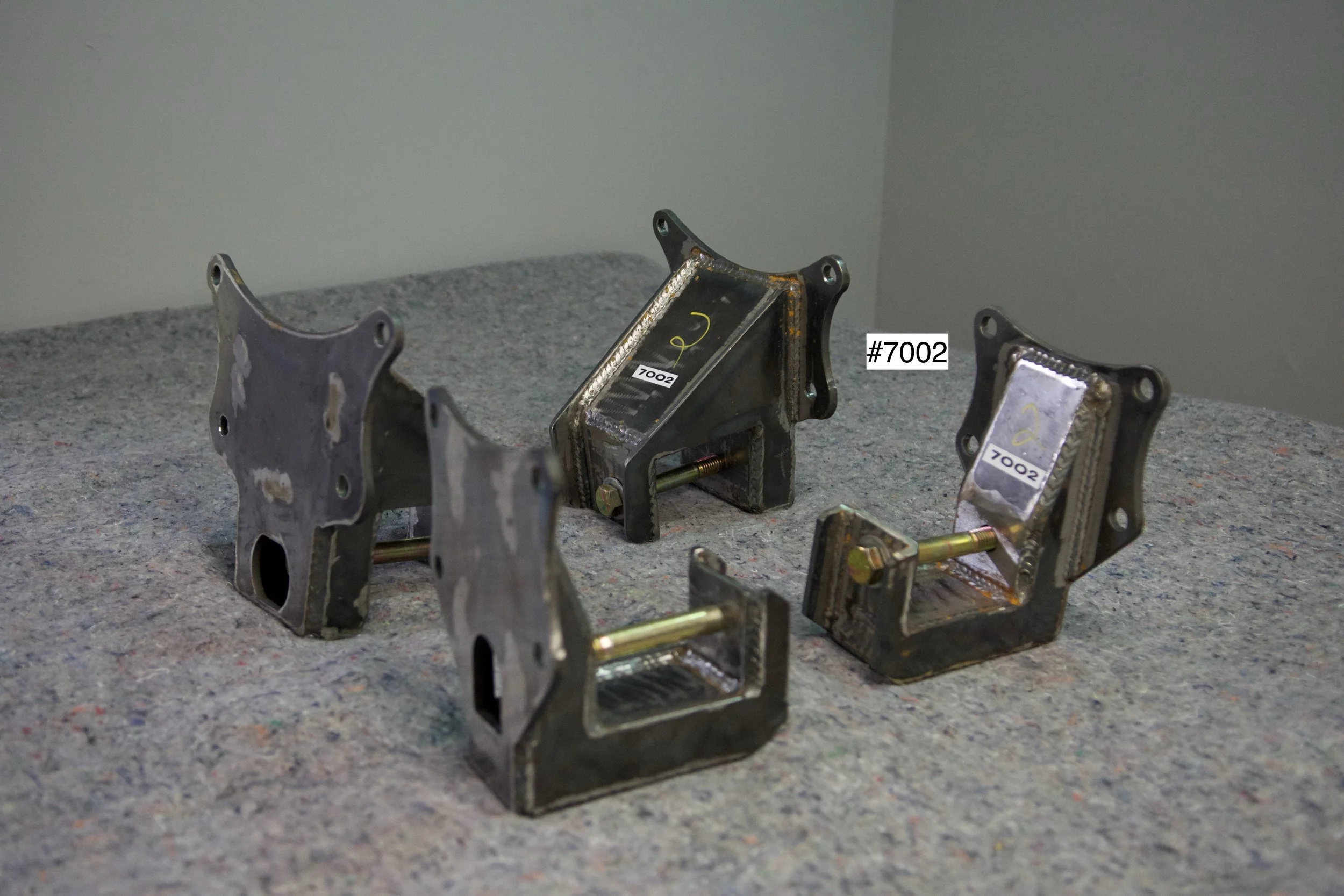 SuperBronco Rear Spring Mounts Rev. 1 – raw bare steel brackets shown from front and side angles, part numbers #7001 and #7002. Fabricated mounts for Bronco suspension builds.