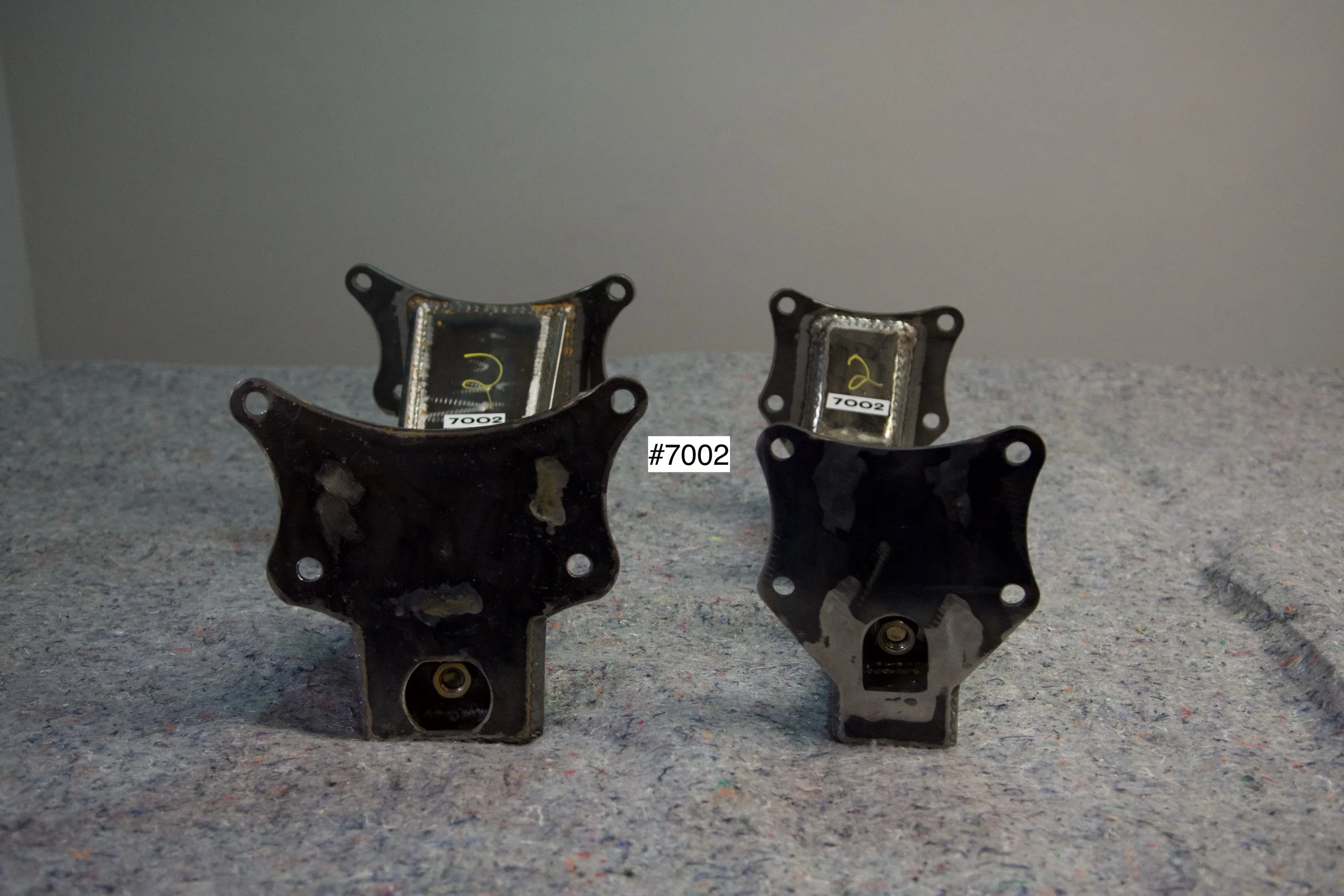 SuperBronco Rear Spring Mounts Rev. 1 – raw bare steel brackets shown from front and side angles, part numbers #7001 and #7002. Fabricated mounts for Bronco suspension builds.