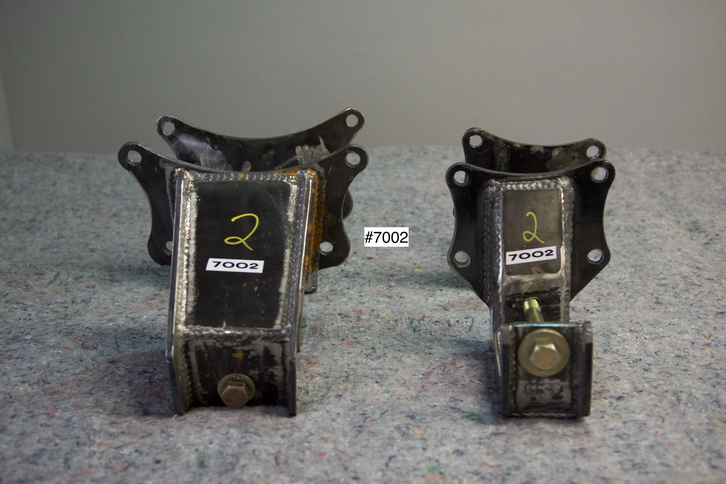 SuperBronco Rear Spring Mounts Rev. 1 – raw bare steel brackets shown from front and side angles, part numbers #7001 and #7002. Fabricated mounts for Bronco suspension builds.