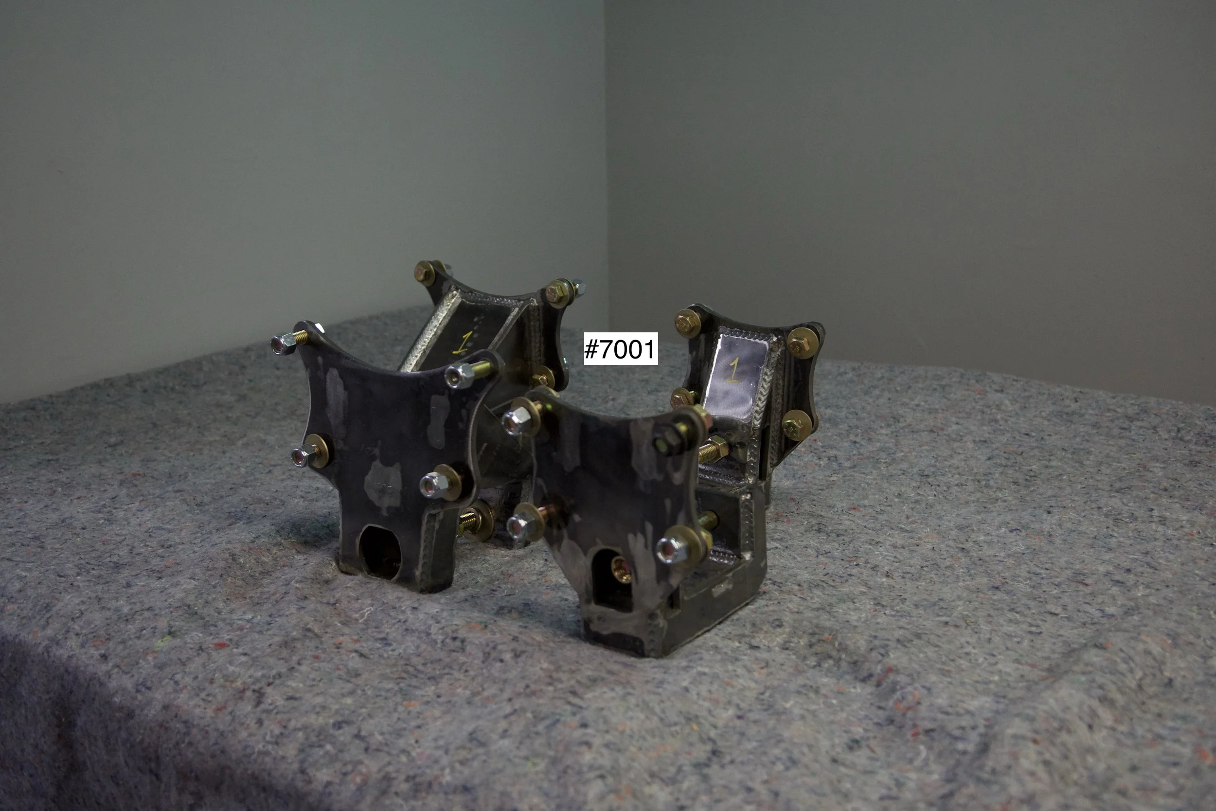SuperBronco Rear Spring Mounts Rev. 1 – raw bare steel brackets shown from front and side angles, part numbers #7001 and #7002. Fabricated mounts for Bronco suspension builds.