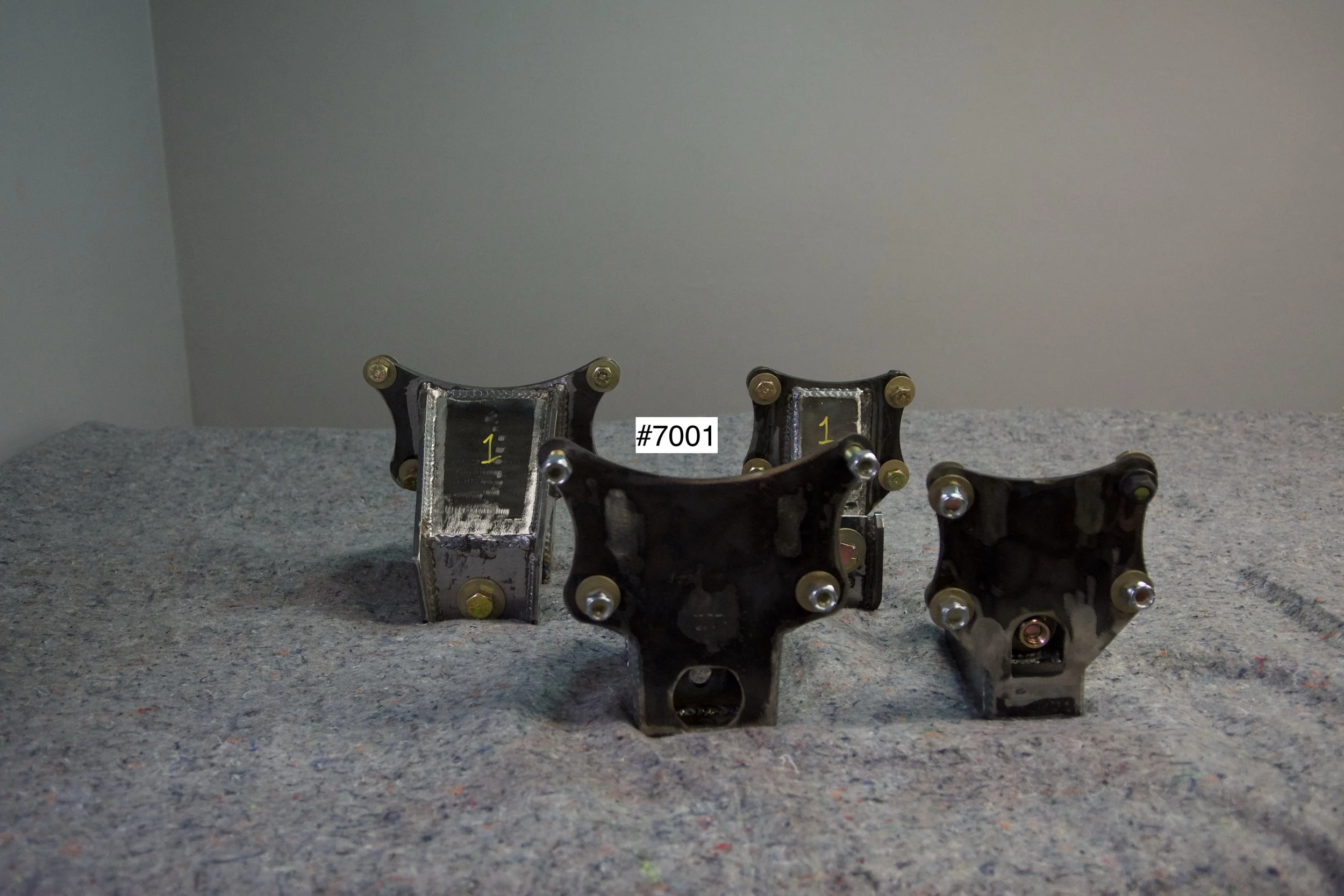 SuperBronco Rear Spring Mounts Rev. 1 – raw bare steel brackets shown from front and side angles, part numbers #7001 and #7002. Fabricated mounts for Bronco suspension builds.